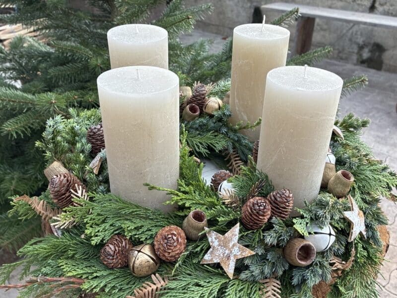 Adventskranz mit vier weißen Kerzen, verziert mit Tannenzweigen, Zapfen und Sternen, draußen aufgestellt.