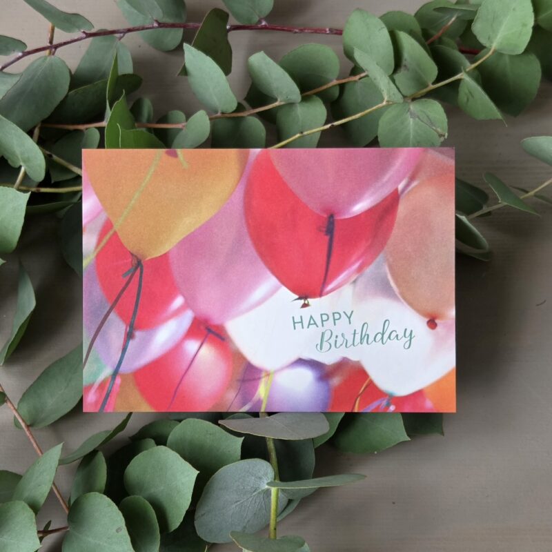 Geburtstagskarte mit bunten Luftballons und Eukalyptusblättern im Hintergrund. Text: Happy Birthday.