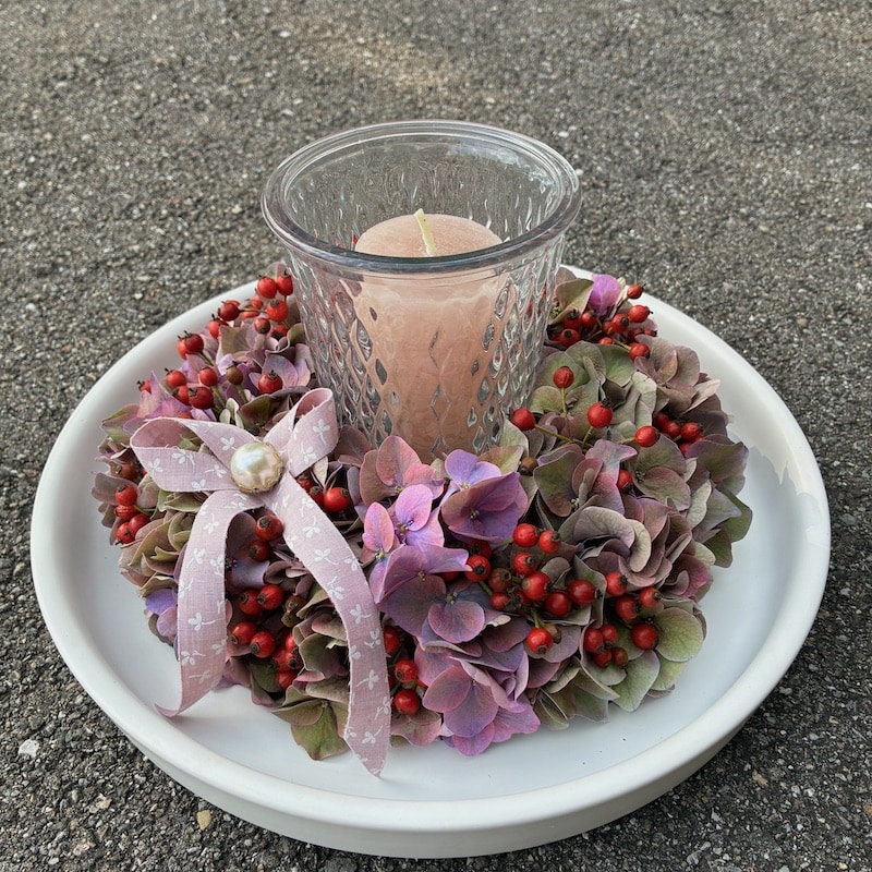 Rosa Kerze im Glas, umgeben von Hortensien und roten Beeren auf einer weißen Schale, dekoriert mit Schleife.